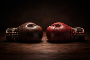Zwei alte Lederboxhandschuhe stehen sich auf einem dunklen Holztisch gegenüber – einer dunkelbraun, einer dunkelrot. Beide sind gleich groß und leicht abgenutzt. Die Handschuhe berühren sich fast in der Mitte, ohne sich zu berühren. Dramatisches Licht von oben, dunkler Hintergrund, starke Schatten. Kein Mensch, keine Hände sind zu sehen.